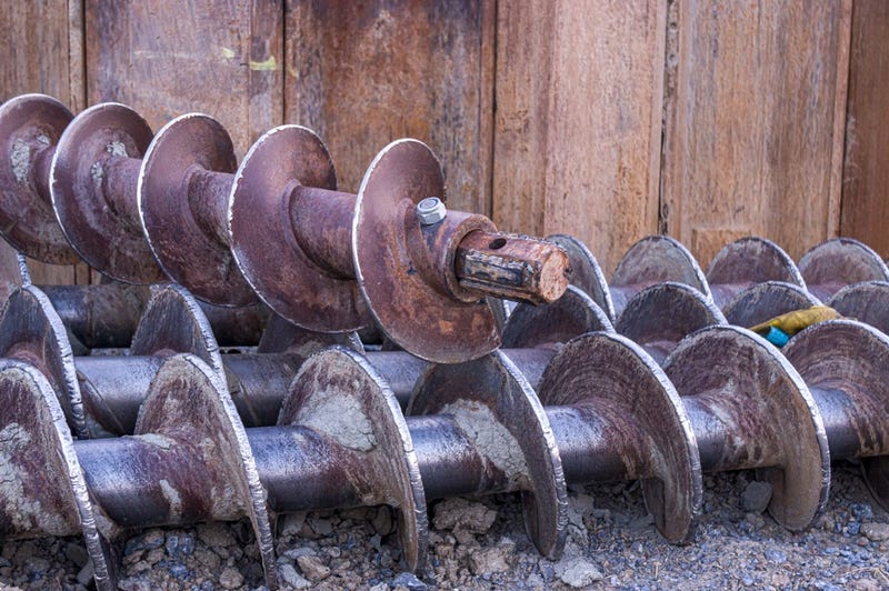 Three earth drills lie side by side on gravel